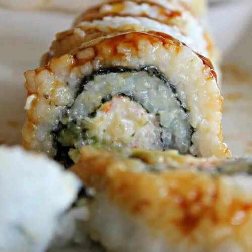 Close-up of sliced sushi roll filled with rice, seaweed, and vegetables, drizzled with sauce, on a white plate.