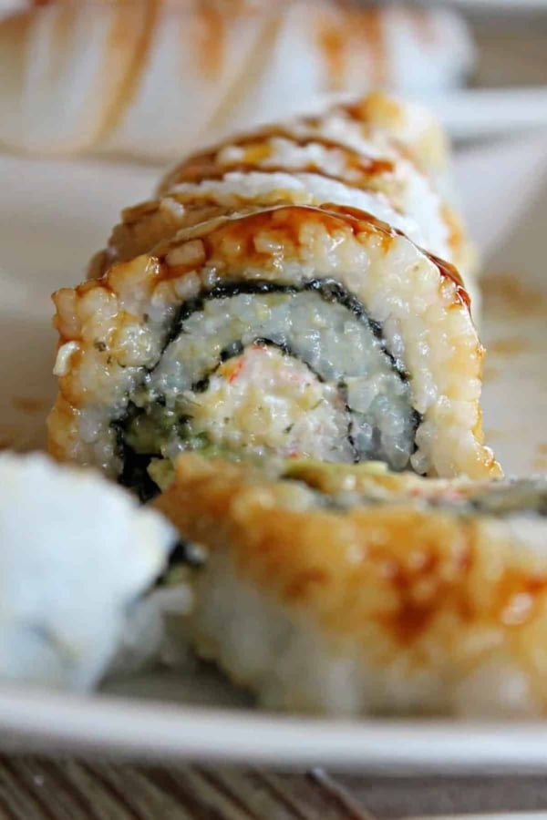 Close-up of sliced sushi roll filled with rice, seaweed, and vegetables, drizzled with sauce, on a white plate.