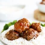 Close-up of a plate with meatballs in sauce, served over white rice with a side of broccoli. A white bowl is in the background.