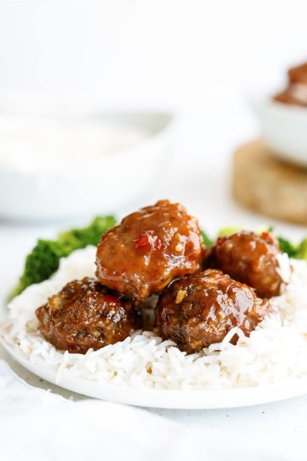 Close-up of a plate with meatballs in sauce, served over white rice with a side of broccoli. A white bowl is in the background.