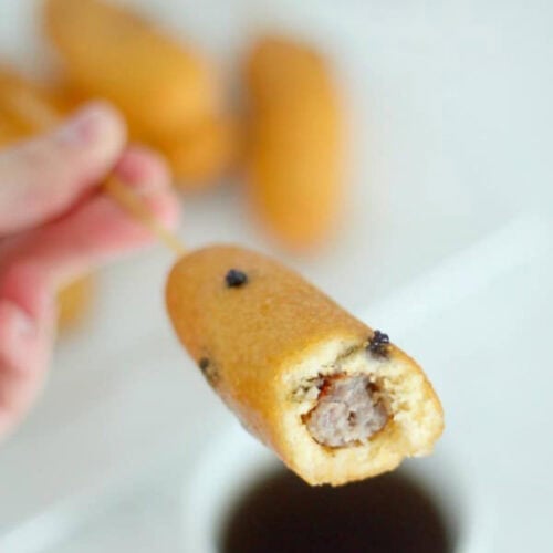A hand holding a corn dog with a bite taken, revealing a sausage inside. Blurred background with more corn dogs and a cup of dipping sauce.