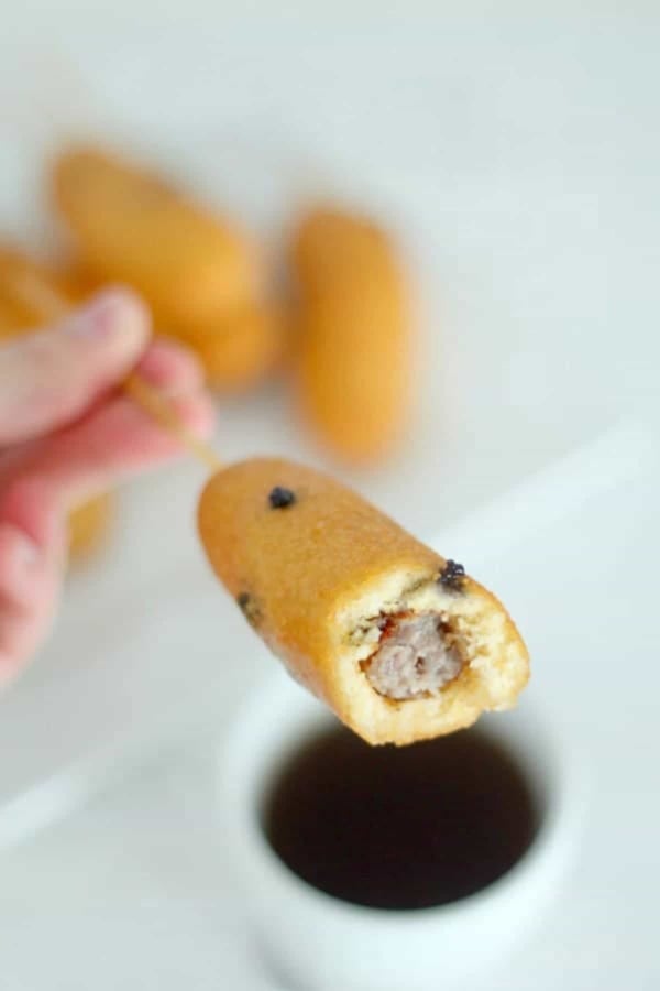 A hand holding a corn dog with a bite taken, revealing a sausage inside. Blurred background with more corn dogs and a cup of dipping sauce.