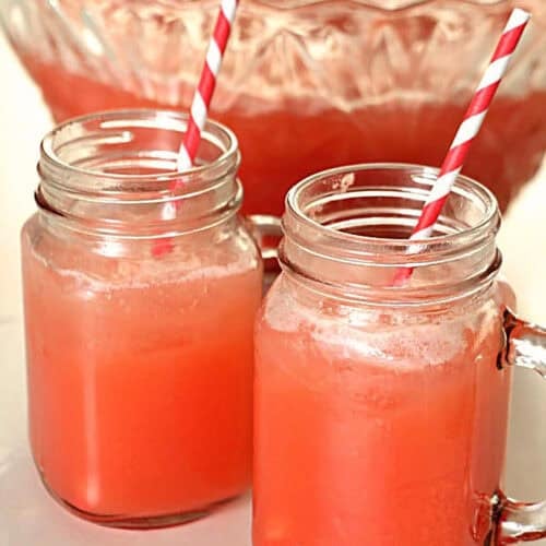 Two mason jar mugs filled with pink punch and striped straws sit in front of a large glass punch bowl with more pink punch.
