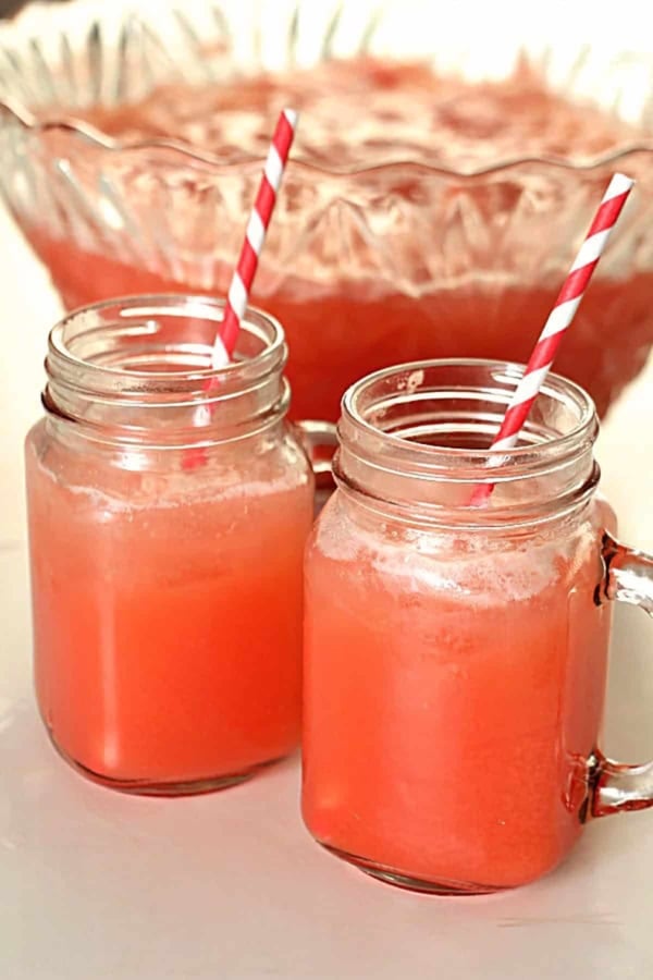 Two mason jar mugs filled with pink punch and striped straws sit in front of a large glass punch bowl with more pink punch.