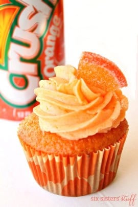 An Orange Crush Cupcake topped with frosting and a small orange slice. A can of Orange Crush in the background.