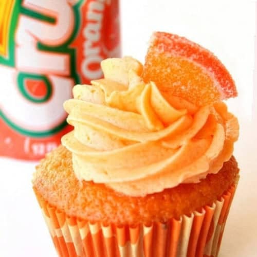 An orange cupcake with swirled frosting and a gummy candy slice on top, placed in front of a can of Orange Crush soda.