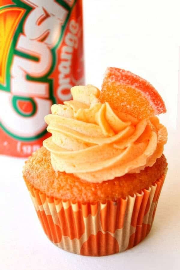 An orange cupcake with swirled frosting and a gummy candy slice on top, placed in front of a can of Orange Crush soda.