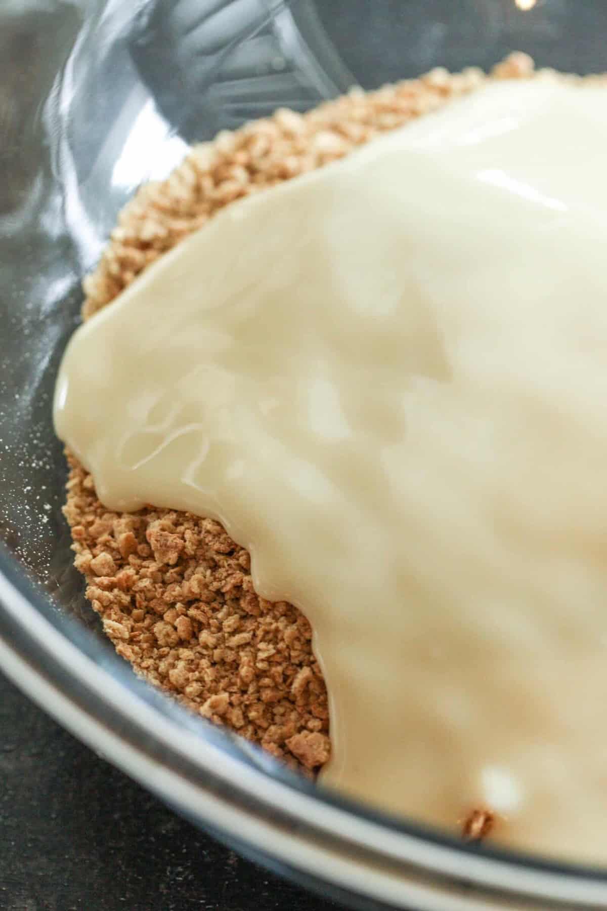 A glass bowl containing crushed graham crackers partially covered with sweetened condensed milk.