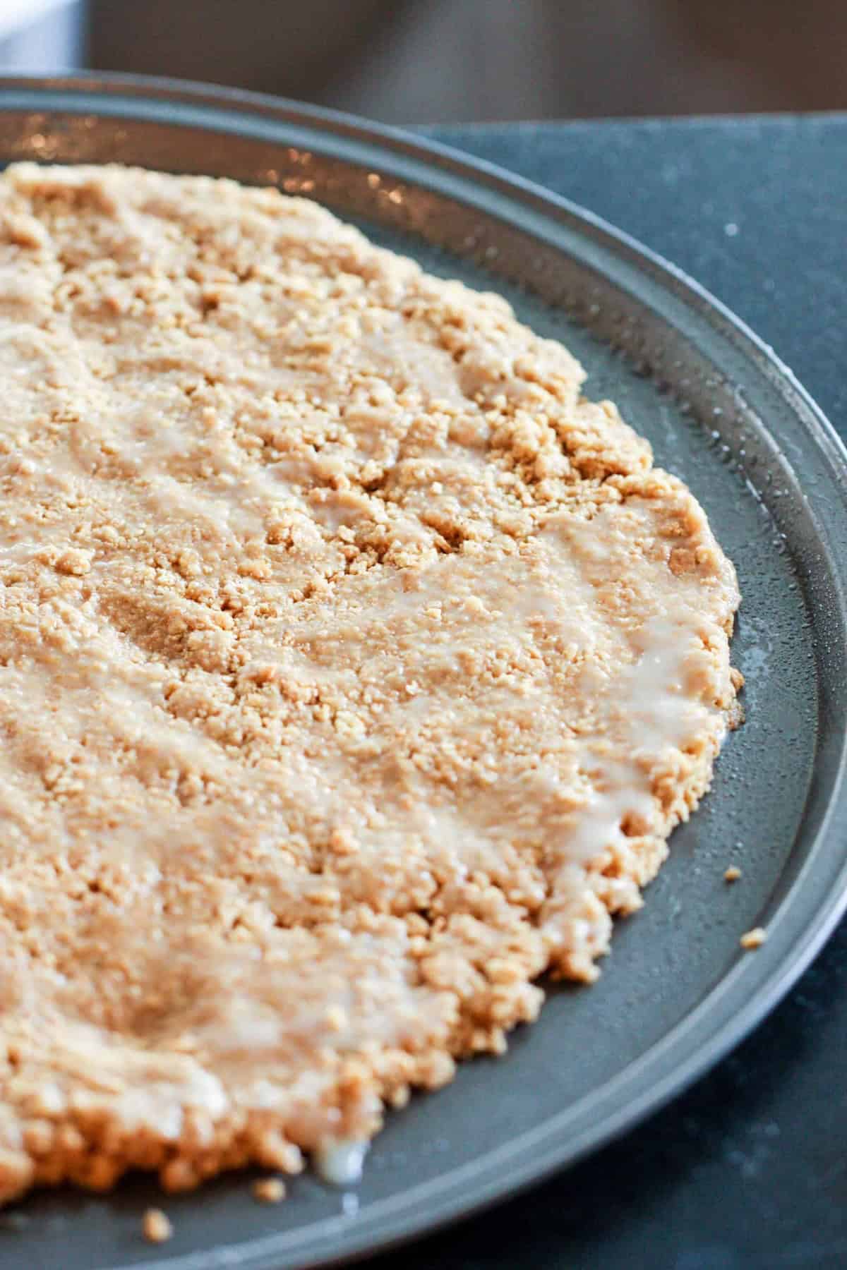 A close-up of an unbaked oatmeal cookie dough pressed into a round metal pizza pan, ready for baking.