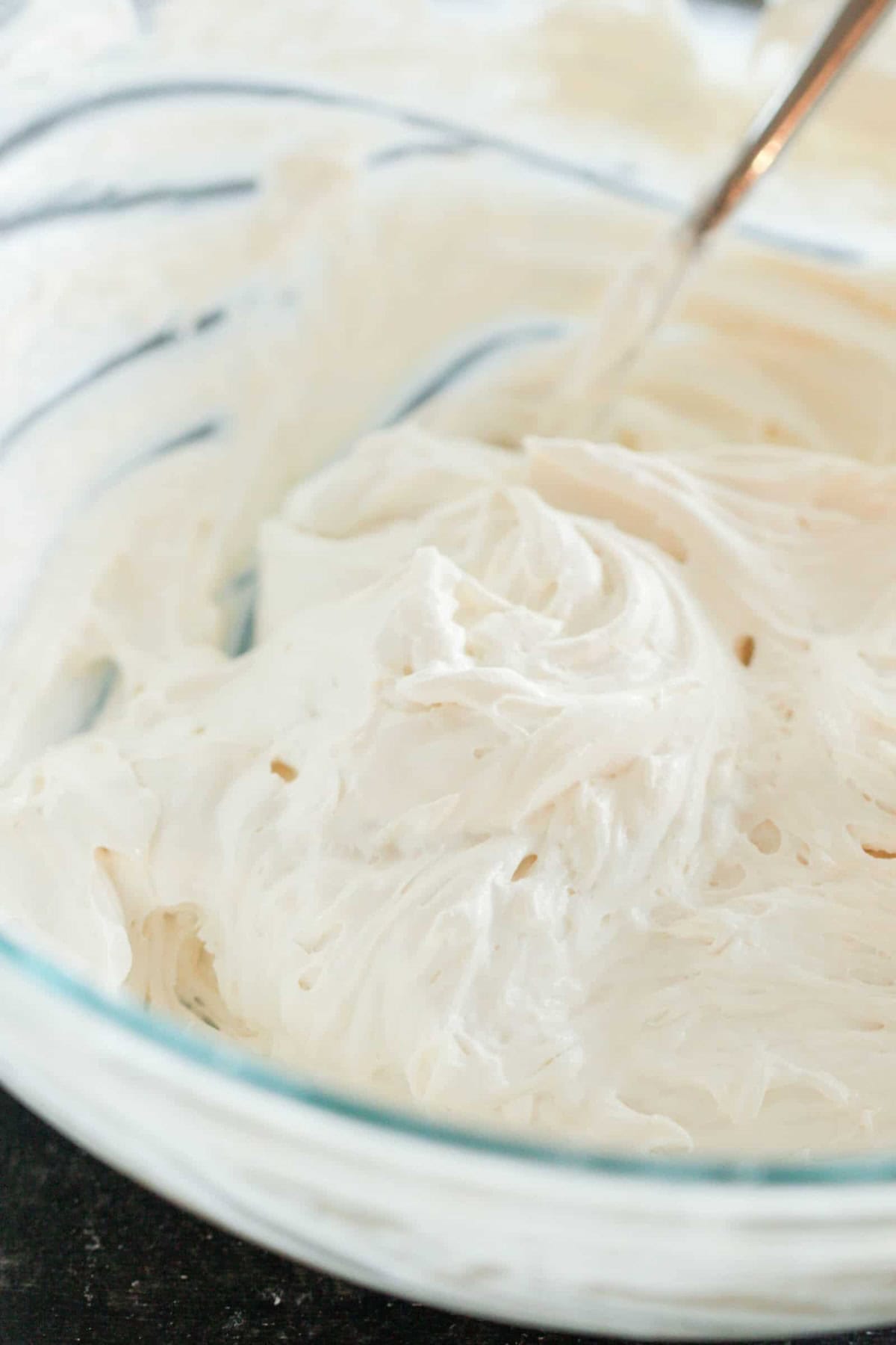 A glass bowl filled with thick, whipped white frosting, with a metal spoon resting inside.