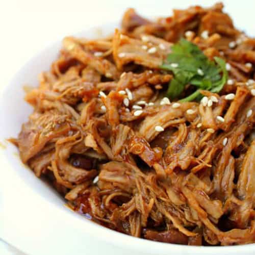 A white bowl filled with shredded, sauced meat garnished with sesame seeds and a sprig of cilantro.