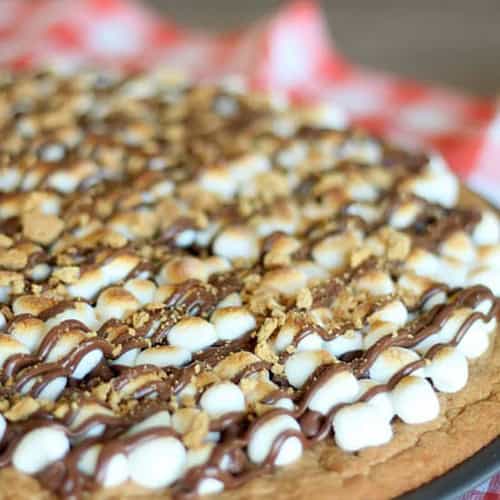 A close-up of a large cookie topped with mini marshmallows, chocolate drizzle, and crushed graham crackers on a red checkered cloth.