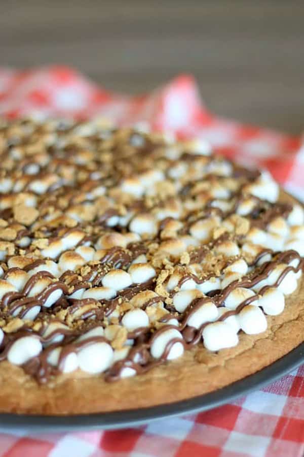 A close-up of a large cookie topped with mini marshmallows, chocolate drizzle, and crushed graham crackers on a red checkered cloth.
