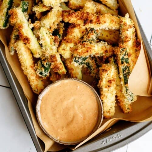 A lined sheet pan crispy zucchini fries and a small cup of dipping sauce.