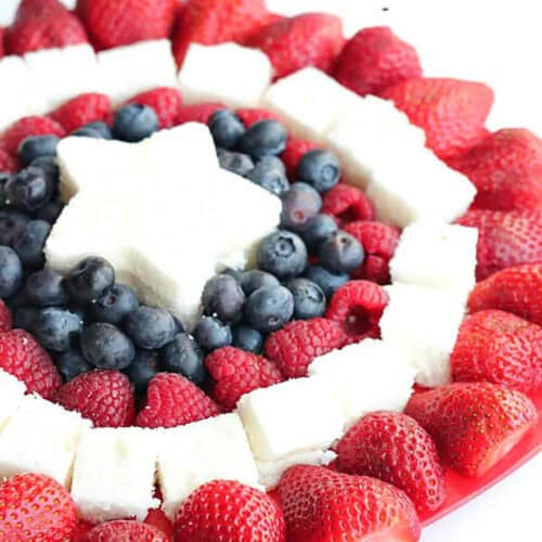A plate arranged with strawberries, raspberries, blueberries, and square pieces of white cake, with a star-shaped cake piece in the center, forming a patriotic pattern.