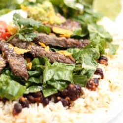 A close-up of a burrito bowl with sliced steak, spinach, rice, black beans, guacamole, and cilantro on a tortilla.