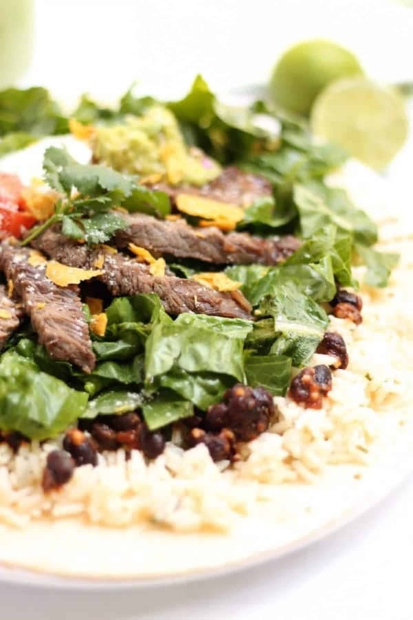 A close-up of a burrito bowl with sliced steak, spinach, rice, black beans, guacamole, and cilantro on a tortilla.