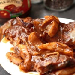 A plate of cooked meat with onions in a brown sauce, with a jar of apple butter and a pepper grinder in the background.
