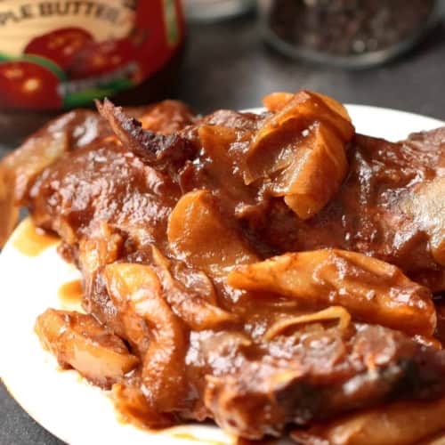 A plate of cooked meat with onions in a brown sauce, with a jar of apple butter and a pepper grinder in the background.