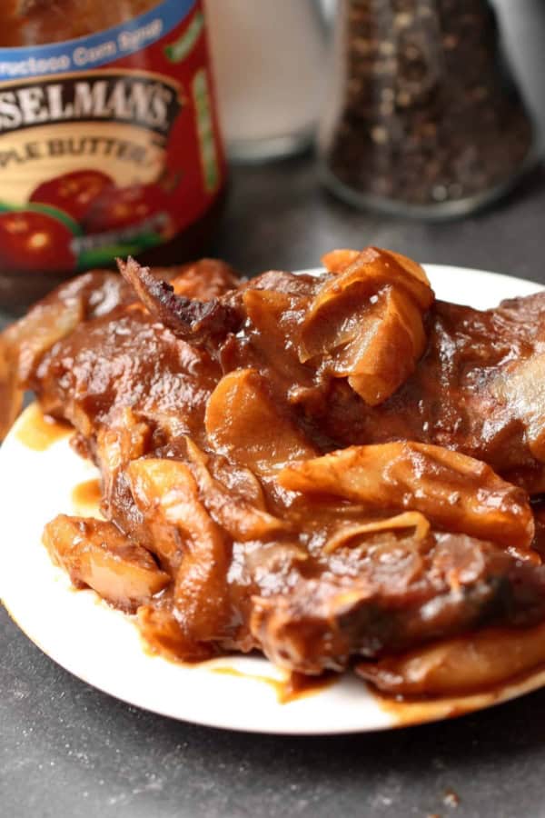 A plate of cooked meat with onions in a brown sauce, with a jar of apple butter and a pepper grinder in the background.