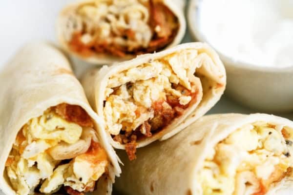Four breakfast burritos filled with eggs, cheese, and other ingredients are displayed on a white plate. A small bowl of sour cream is next to the burritos.