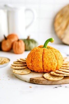 A pumpkin-shaped cheeseball sits on a round wooden board, surrounded by crackers. In the background, there are small pumpkins and a bowl of snacks.