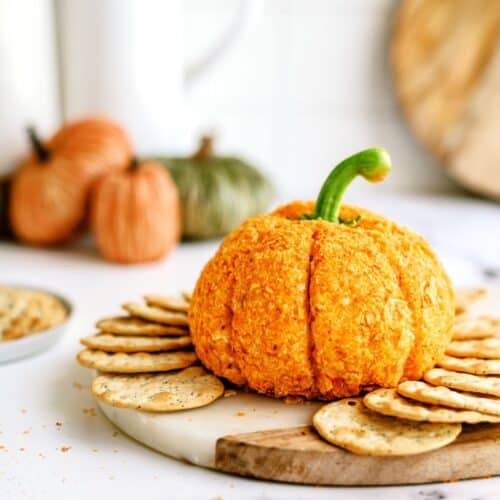 A pumpkin-shaped cheeseball sits on a round wooden board, surrounded by crackers. In the background, there are small pumpkins and a bowl of snacks.