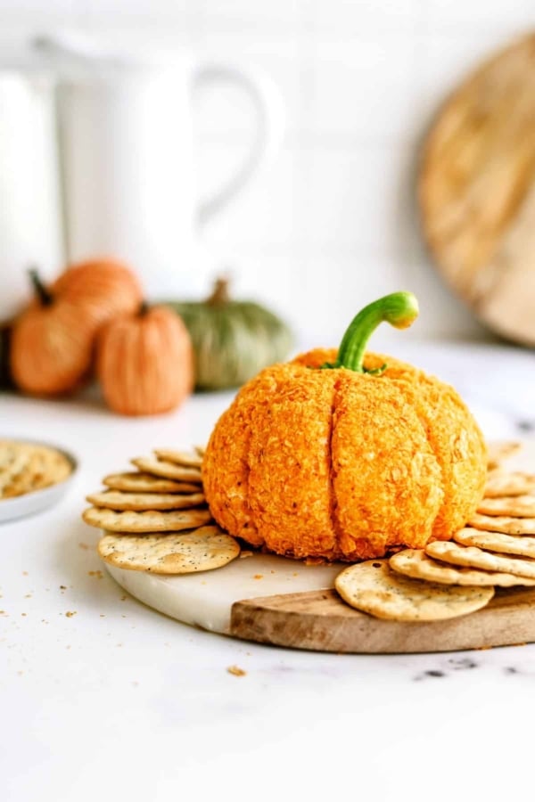 A pumpkin-shaped cheeseball sits on a round wooden board, surrounded by crackers. In the background, there are small pumpkins and a bowl of snacks.