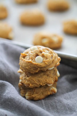A stack of four freshly baked cookies with white chocolate chips is placed on a gray cloth, while additional cookies are seen on a baking sheet in the background.