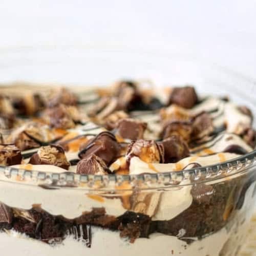 A glass bowl filled with layered dessert, featuring whipped cream, chocolate pieces, brownie chunks, and caramel drizzle.