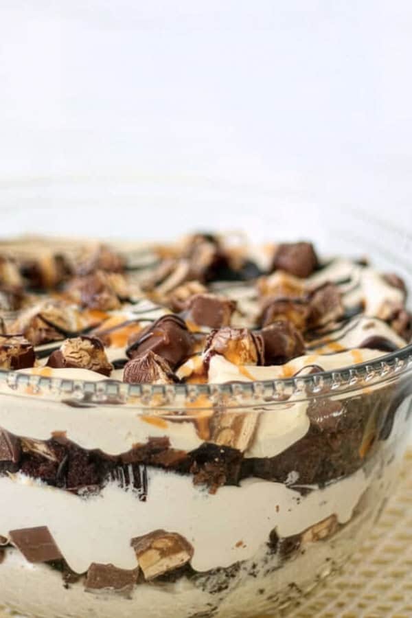 A glass bowl filled with layered dessert, featuring whipped cream, chocolate pieces, brownie chunks, and caramel drizzle.