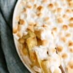 A close-up of a baked dish with a golden-brown marshmallow topping and a serving spoon scooping out a portion, revealing contents underneath.