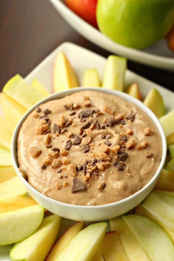 A bowl of creamy dip garnished with crushed nuts and chocolate chips is surrounded by sliced apple wedges.
