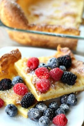 Two squares of German Pancakes on a plate finished with powdered sugar with berries. Pan of remaining German Pancakes in the background.
