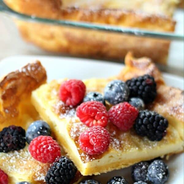Two squares of German Pancakes on a plate finished with powdered sugar with berries. Pan of remaining German Pancakes in the background.