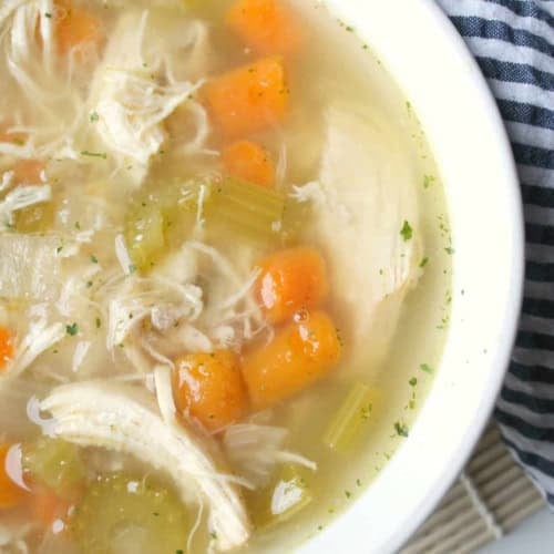 A bowl of chicken soup with shredded chicken, diced carrots, celery, and herbs, placed on a striped cloth and bamboo mat.