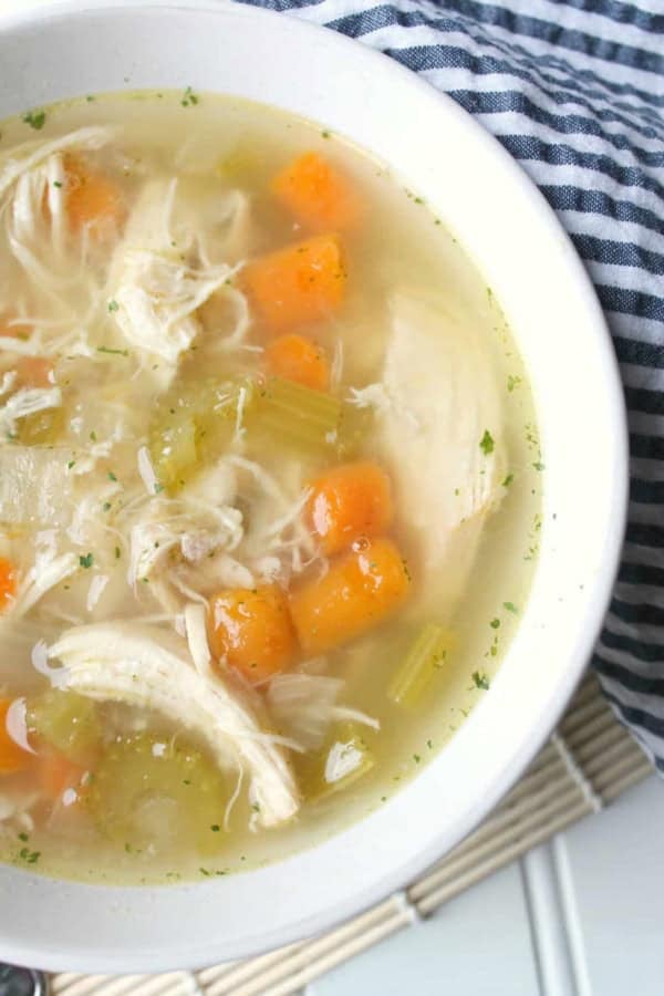 A bowl of chicken soup with shredded chicken, diced carrots, celery, and herbs, placed on a striped cloth and bamboo mat.