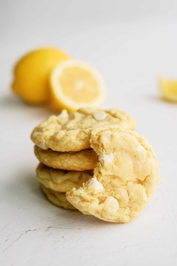Lemon Cheesecake Pudding Cookies stacked with on cookie leaning on the stack missing a bite. Fresh lemons in the background.
