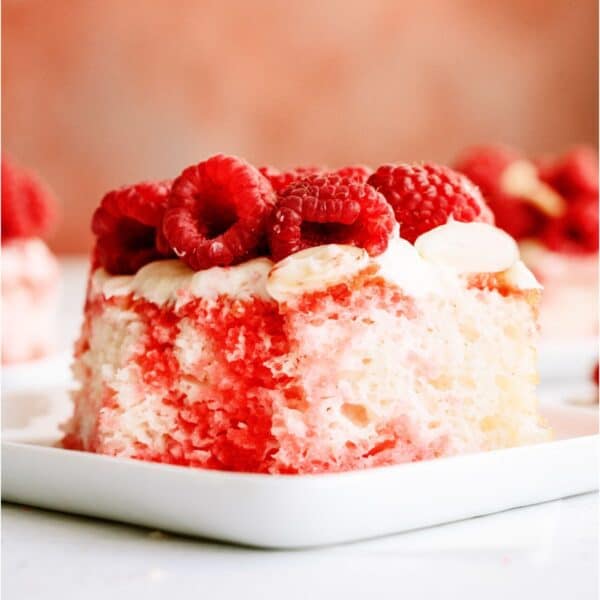 A slice of White Raspberry Poke Cake on a white plate topped with fresh raspberries and chopped almonds. More slices on plates in the background.