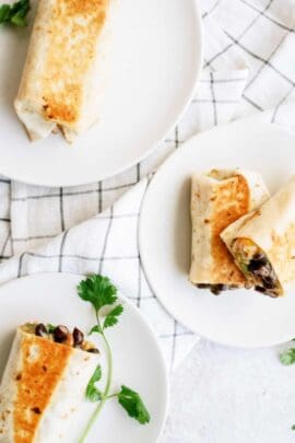 Three grilled burritos on white plates with a sprig of cilantro, placed on a checkered cloth background.