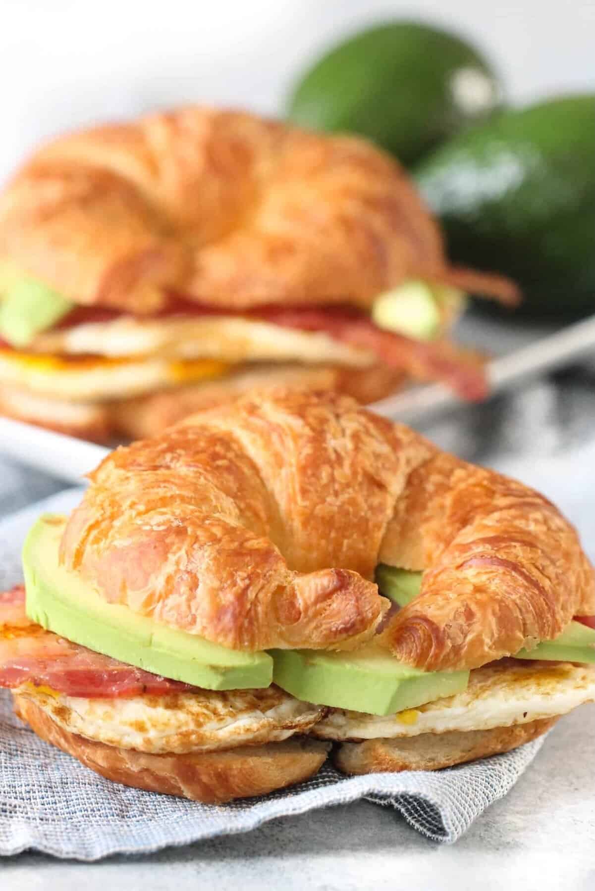Croissant sandwiches with avocado, egg, and bacon on a cloth napkin, with two whole avocados in the background.