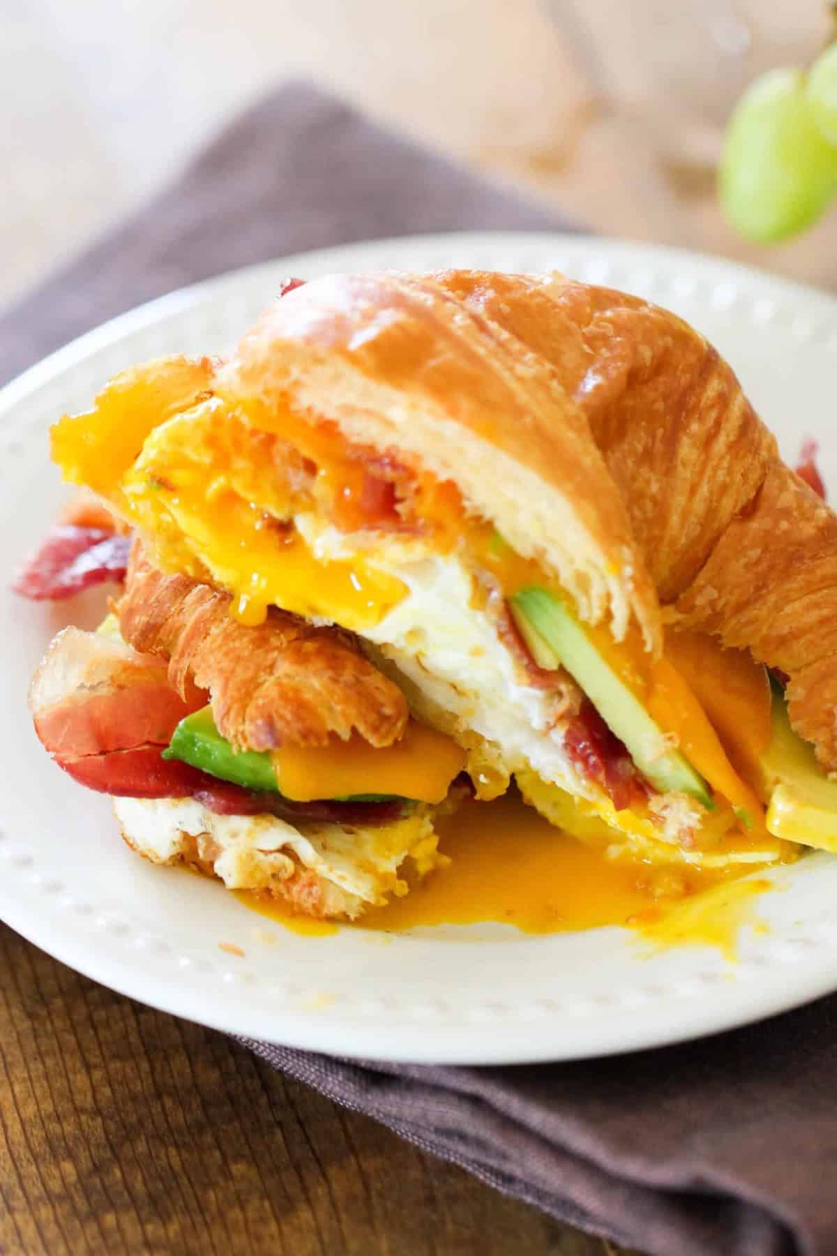A croissant sandwich with egg, cheese, bacon, and avocado on a white plate.