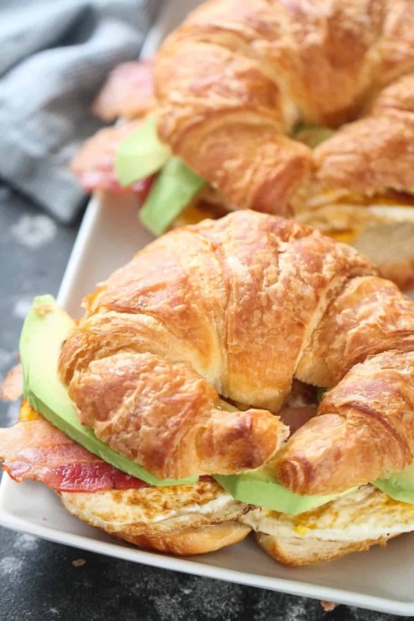 Two croissant sandwiches with avocado, egg, and bacon on a white plate.