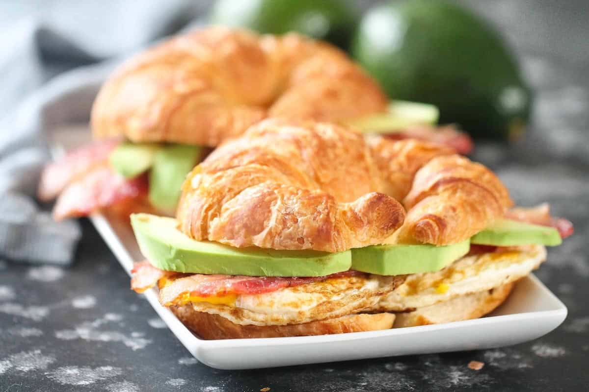Two croissant sandwiches with avocado, egg, and bacon on a rectangular white plate. Two avocados in the background.