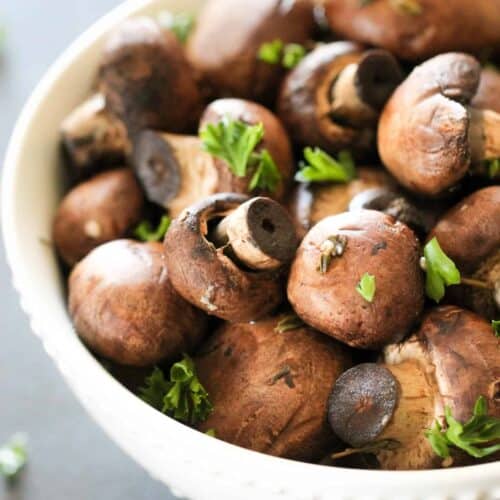 A white bowl filled with whole brown mushrooms, garnished with fresh parsley, placed on a dark surface.