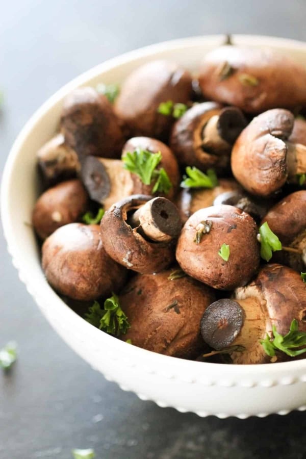 A white bowl filled with whole brown mushrooms, garnished with fresh parsley, placed on a dark surface.