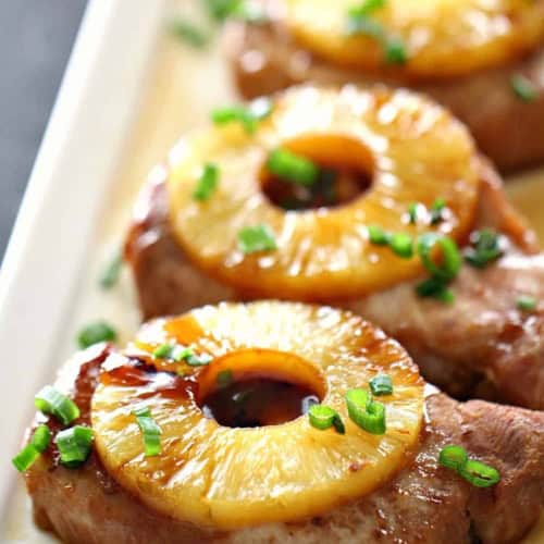 Pork chops topped with pineapple rings and chopped green onions arranged on a white rectangular plate.