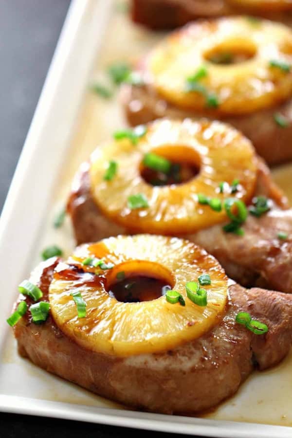 Pork chops topped with pineapple rings and chopped green onions arranged on a white rectangular plate.