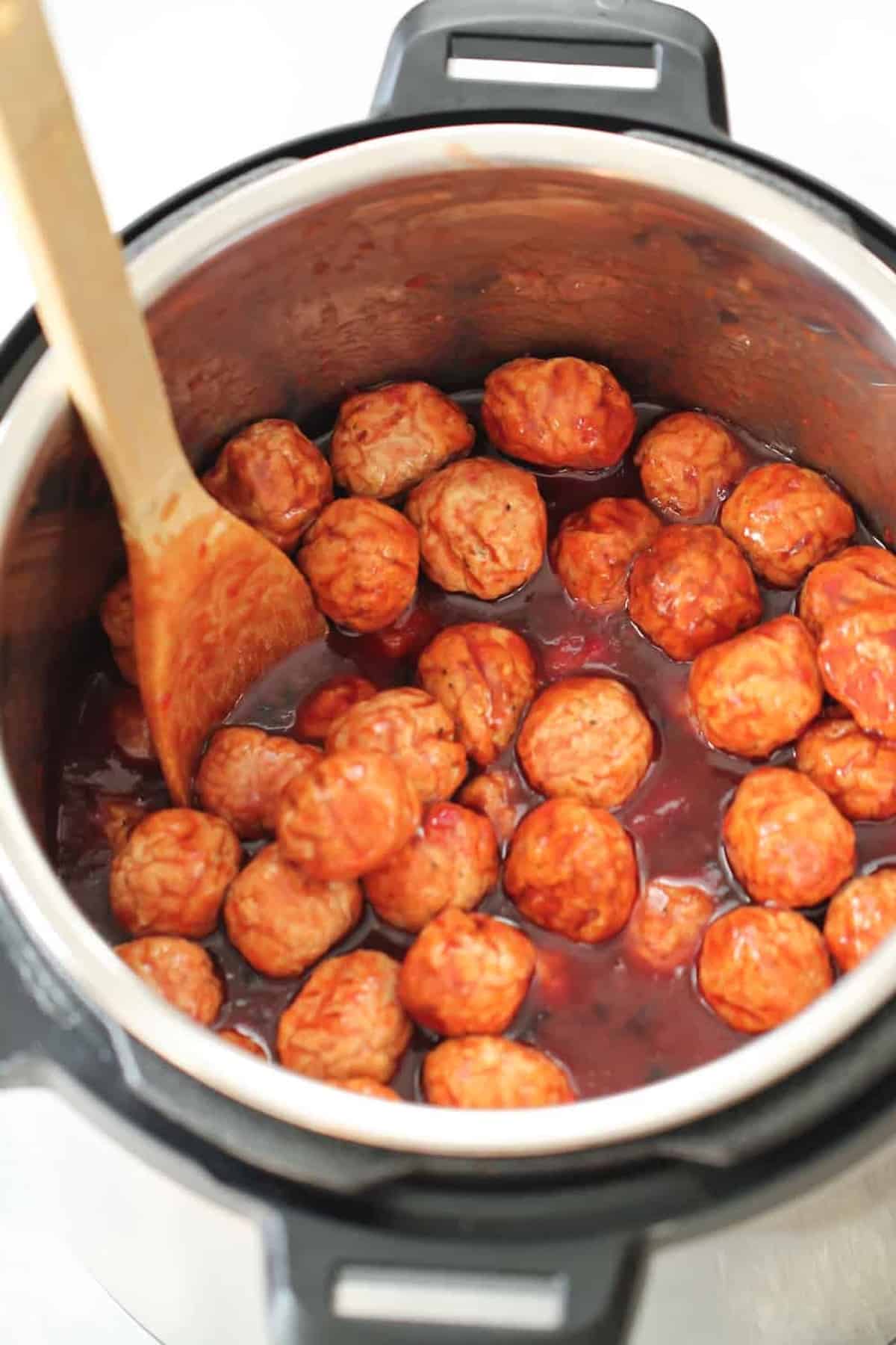 Meatballs cooking in a sauce inside an Instant Pot with a wooden spoon.