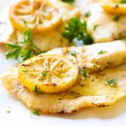 Cooked fish fillets garnished with slices of lemon and chopped parsley on a white plate.